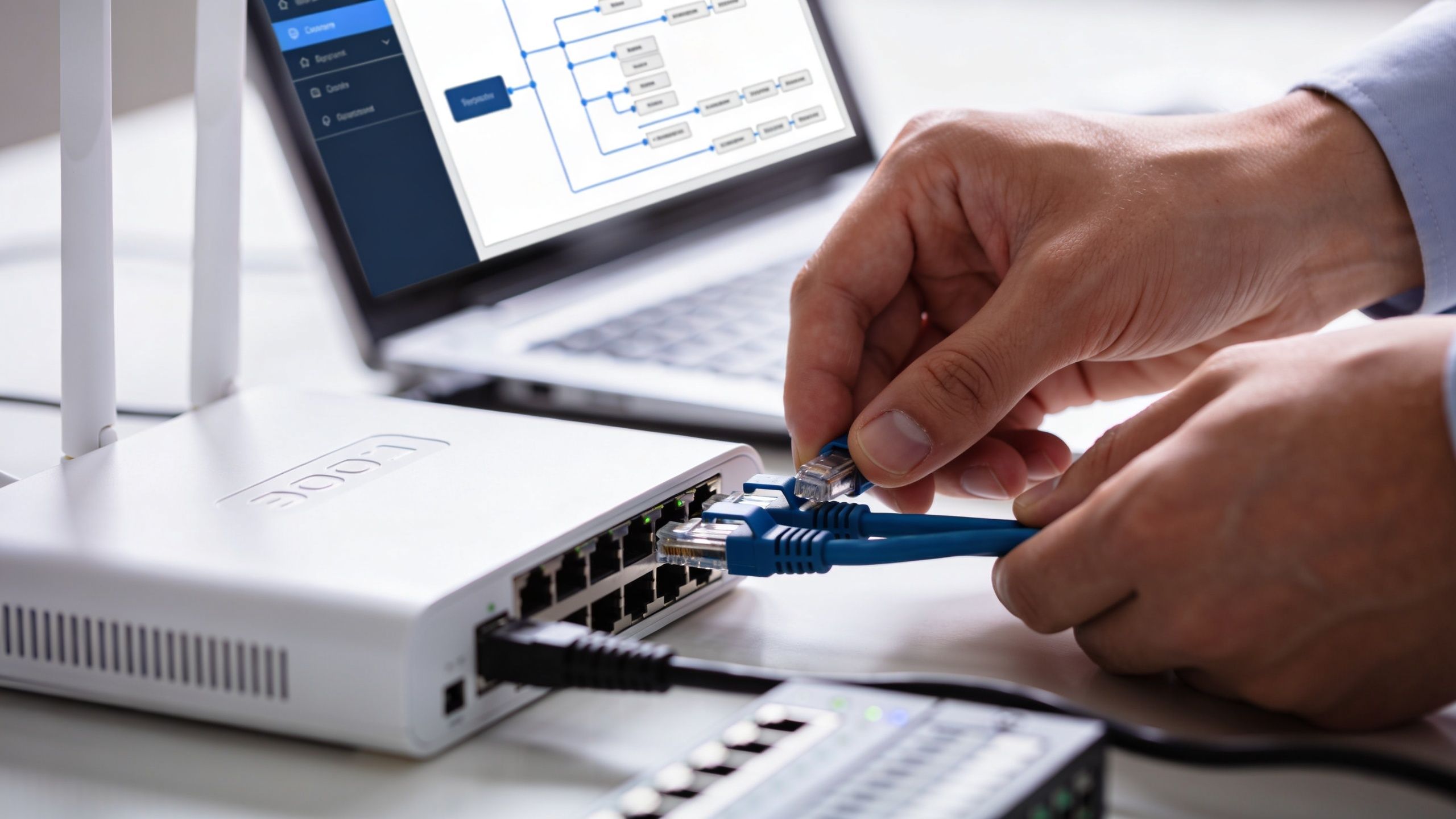 Ein Techniker verbindet ein blaues Netzwerkkabel mit einem Netzwerk-Switch, während ein Laptop im Hintergrund Diagramme anzeigt.
